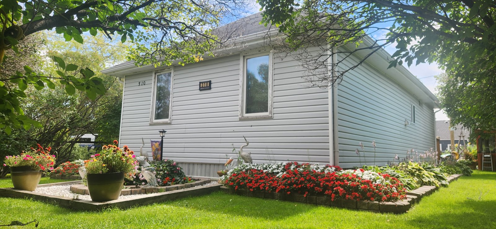 310 Epsilon Avenue, Winnipeg Beach &middot; 310 Epsilon Avenue seen under mature trees, with flower borders running along the house